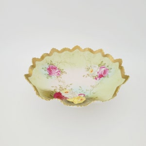 May include: A white porcelain bowl with a scalloped edge and a gold rim. The bowl is decorated with a floral pattern of pink, red, and yellow roses on a green background.