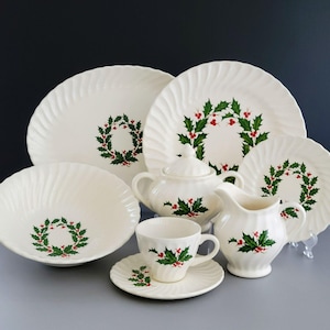 May include: A cream-colored Christmas dinnerware set featuring a holly wreath design. The set includes plates, a bowl, a sugar bowl, a creamer, a teacup, and a saucer. The plates and bowl have a swirled rim design.