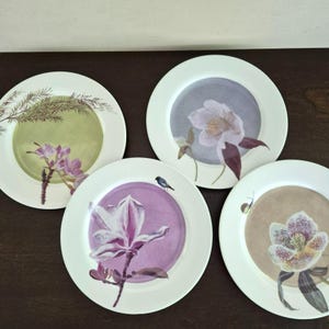 May include: Four round white ceramic plates featuring distinct botanical illustrations. One plate shows pink flowers on a green background, another a purple flower, a third a white flower on a gray background, and the last a spotted flower with a snail.