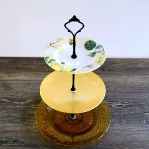 May include: A three-tiered serving stand with a black metal frame. The top tier features a white plate with a floral design, the middle tier is a solid yellow plate, and the bottom tier is a textured amber-colored plate.