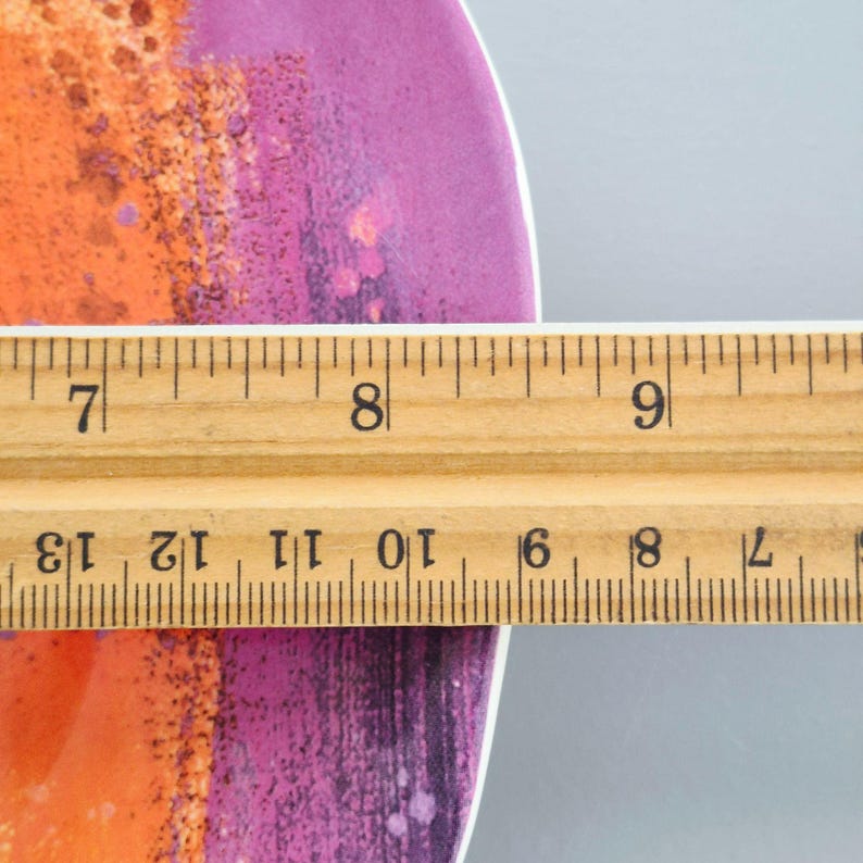 May include: A ceramic plate with an abstract design in shades of orange and purple. A wooden ruler is placed across the plate, showing measurements in inches. The plate's design has a textured appearance.