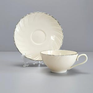 May include: Cream-colored teacup and saucer set with a silver trim. The saucer has a spiral design, and the teacup has a handle. The set is displayed on a light gray surface, showcasing a classic and elegant design.