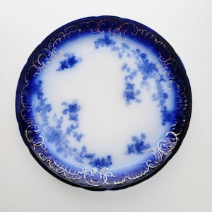 May include: A round, antique porcelain plate with a white center and a deep blue rim. The rim features gold filigree and a floral pattern in blue. The plate's design is a classic example of vintage tableware.
