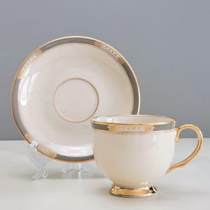 May include: Cream-colored teacup and saucer set with gold trim and a gray border. The teacup has a gold handle and base. The saucer has a matching gray border and gold accents. The set is displayed on a clear stand.