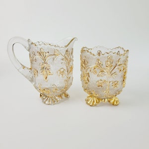 May include: A clear glass creamer and sugar bowl set. Both pieces feature a textured surface and are adorned with gold floral and leaf designs. The creamer has a handle and spout, while the sugar bowl has a scalloped rim and sits on a gold pedestal base.