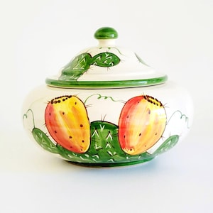 May include: A white ceramic jar with a lid, hand-painted with green cacti and yellow-orange prickly pear fruit. The lid has a green knob and a green border. The jar is a decorative kitchen item.