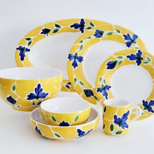 May include: A collection of yellow ceramic tableware with a floral design. The set includes plates, bowls, and a mug, all adorned with blue flowers and green leaves. The tableware has a bright, cheerful aesthetic.