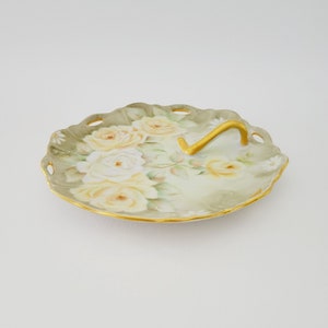 May include: Oval-shaped porcelain serving dish with a gold handle and rim. The dish features a hand-painted design of yellow roses and green leaves on a pale green background. The edges have a scalloped design.