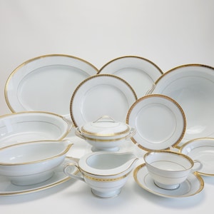 May include: A set of white porcelain dinnerware with gold trim. The set includes a large oval platter, a round serving bowl, a gravy boat, a creamer, a sugar bowl, a teacup and saucer, and several dinner plates.