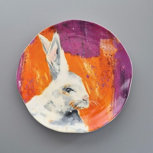 May include: A ceramic plate with a painted design of a white rabbit. The background is a mix of orange and purple hues, with the rabbit's portrait in the center. The plate is a decorative item, suitable for display or serving.