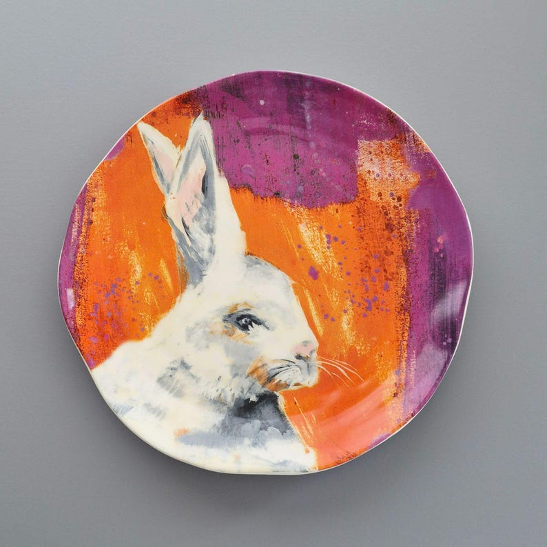 May include: A ceramic plate with a painted design of a white rabbit. The background is a mix of orange and purple hues, with the rabbit's portrait in the center. The plate is a decorative item, suitable for display or serving.