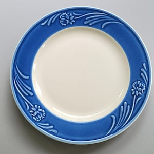 May include: A round, white ceramic plate with a wide, cobalt blue border. The border features a repeating floral and leaf design. The plate's center is a creamy white color, creating a contrast with the blue rim. This decorative plate is suitable for serving food.