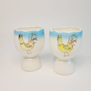 May include: Two white ceramic egg cups with a blue and white design. Each cup features a rooster with a yellow body, green head, and brown tail feathers.