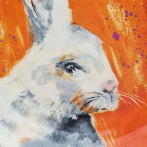 May include: A detailed illustration of a white rabbit's head against a bright orange background. The rabbit features gray and orange shading, a pink nose, and purple paint splatters. The artwork has a painterly style.