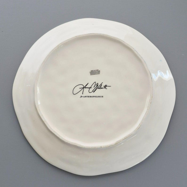May include: White ceramic plate with a wavy edge. The underside features a signature and the text "for ANTHROPOLOGIE". The plate has a glossy finish.