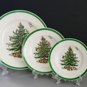 May include: Set of three white ceramic plates with a green border. Each plate features a Christmas tree design with ornaments and gifts. The plates are displayed on clear plastic stands, showcasing a festive holiday theme.