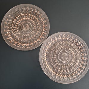 K&ouml;nnte beinhalten: Zwei klare, kreisf&ouml;rmige Teller mit einem detaillierten, geometrischen Muster. Die Teller haben ein erhabenes, strukturiertes Design, das von der Mitte ausgeht. Die Teller sind aus Glas und haben einen rosafarbenen Farbton. Die Teller werden auf dunklem Hintergrund pr&auml;sentiert.