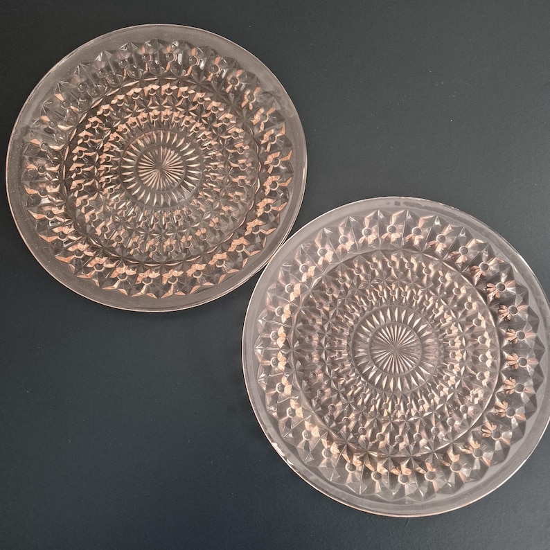 K&ouml;nnte beinhalten: Zwei klare, kreisf&ouml;rmige Teller mit einem detaillierten, geometrischen Muster. Die Teller haben ein erhabenes, strukturiertes Design, das von der Mitte ausgeht. Die Teller sind aus Glas und haben einen rosafarbenen Farbton. Die Teller werden auf dunklem Hintergrund pr&auml;sentiert.