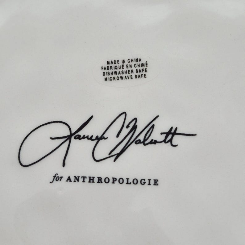 May include: White ceramic dish with black text and signature. The text includes "Made in China", "Dishwasher Safe", "Microwave Safe", and "for ANTHROPOLOGIE". The signature is in black cursive.