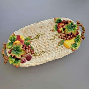 May include: Oval ceramic serving tray with a woven basket design. The tray is decorated with colorful, three-dimensional fruit including grapes, peaches, and pears. The handles are designed to look like tree branches.