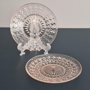 K&ouml;nnte beinhalten: Zwei rosa Glasplatten mit Rautenmuster. Eine Platte wird auf einem transparenten Kunststoffst&auml;nder pr&auml;sentiert. Die Platten haben ein kreisf&ouml;rmiges Design mit einem Sternmittelpunkt. Die Platten befinden sich auf einer dunkelblauen Oberfl&auml;che.