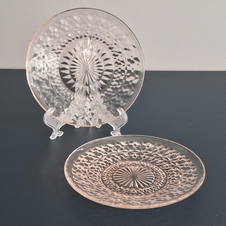 K&ouml;nnte beinhalten: Zwei rosa Glasplatten mit Rautenmuster. Eine Platte wird auf einem transparenten Kunststoffst&auml;nder pr&auml;sentiert. Die Platten haben ein kreisf&ouml;rmiges Design mit einem Sternmittelpunkt. Die Platten befinden sich auf einer dunkelblauen Oberfl&auml;che.