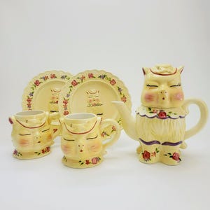 May include: A vintage-style tea set featuring a teapot, two mugs, and two plates. The set is primarily yellow with a cat face design, floral accents, and a scalloped edge. The teapot has a long spout and a handle.