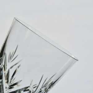 K&ouml;nnte beinhalten: Nahaufnahme eines klaren, geschliffenen Kristallglases. Das Glas hat ein geometrisches Muster aus Linien und Formen, mit einer leicht abgewinkelten Ansicht. Das Glas ist leer und vor einem wei&szlig;en Hintergrund platziert.