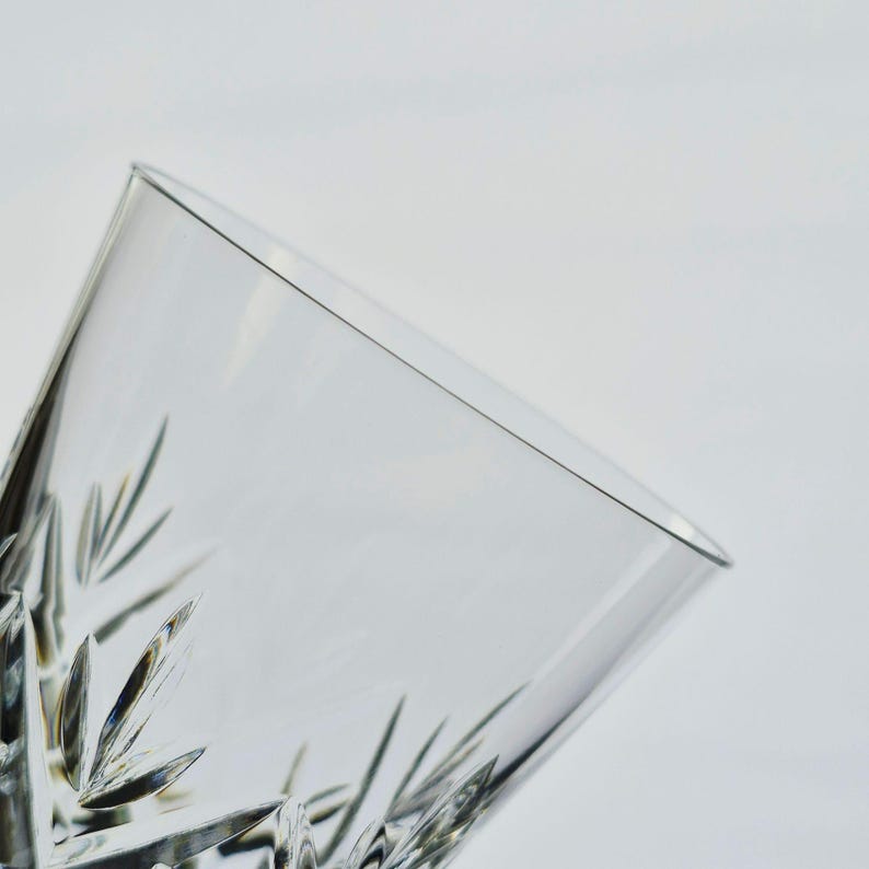 K&ouml;nnte beinhalten: Nahaufnahme eines klaren, geschliffenen Kristallglases. Das Glas hat ein geometrisches Muster aus Linien und Formen, mit einer leicht abgewinkelten Ansicht. Das Glas ist leer und vor einem wei&szlig;en Hintergrund platziert.