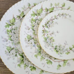 May include: A set of white porcelain plates with a floral design. The plates feature a delicate pattern of white flowers and green leaves, with gold trim around the edges. The plates are arranged in a nested configuration.