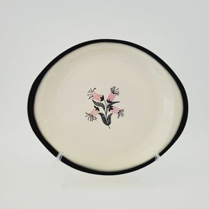 May include: A cream-colored plate with a black rim and a floral design. The flowers are pink with black details, and the stems are black. The plate has an irregular, slightly curved shape.