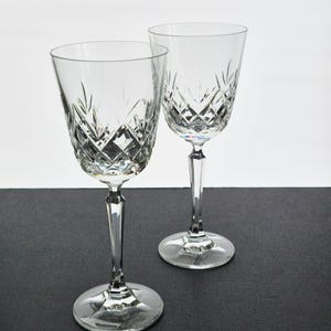 K&ouml;nnte beinhalten: Zwei klare Kristall-Weingl&auml;ser mit Diamant- und vertikalen Linienmustern. Die Gl&auml;ser haben lange Stiele und runde Basen. Der Hintergrund ist ein neutrales Grau und Wei&szlig;.