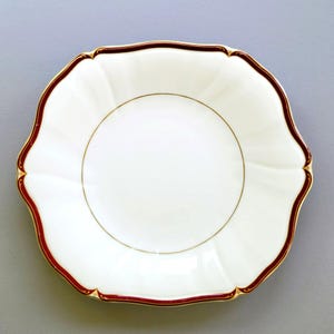 May include: A white scalloped-edge plate with a burgundy border and gold trim. The plate has a gold inner ring and a simple, elegant design. The plate is likely made of porcelain or fine china.