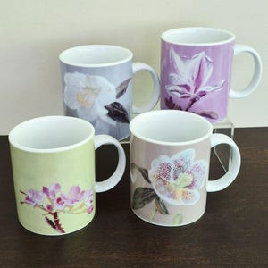 May include: Four ceramic mugs with white interiors and handles, each featuring a unique floral illustration. The designs include a white flower on a gray background, a purple magnolia, pink blossoms on a light green base, and a speckled flower on tan.