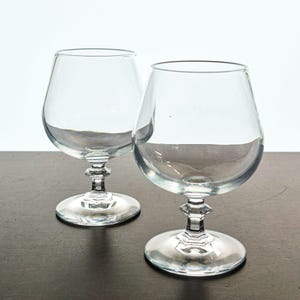 May include: Two clear glass snifters with wide, rounded bowls and short, faceted stems. Each glass features a circular base and a tapered rim, designed for serving spirits. The glassware is transparent and smooth, showcasing a classic silhouette.