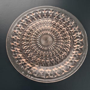 K&ouml;nnte beinhalten: Ein klarer, runder Glasteller mit einem detaillierten, strukturierten Muster. Das Design zeigt ein zentrales Sonnenstrahlmotiv, umgeben von konzentrischen Ringen aus rautenf&ouml;rmigen und geometrischen Elementen. Der Rand des Tellers ist leicht erh&ouml;ht.