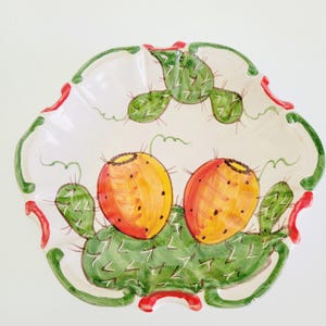 May include: A white ceramic serving dish with a scalloped edge, featuring a hand-painted design of green cacti and two orange and yellow prickly pears. The rim is decorated with green and red accents.