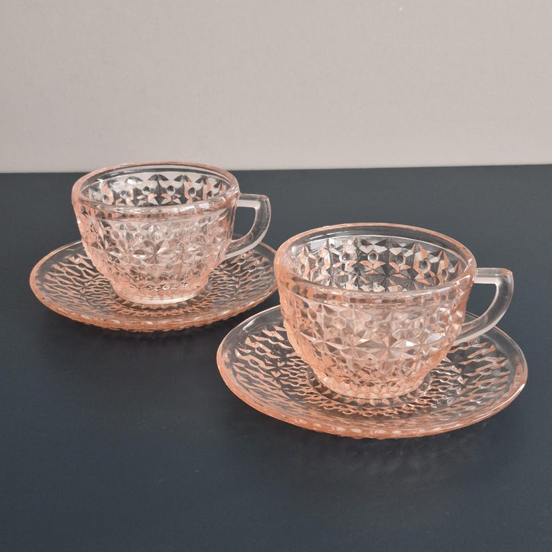 K&ouml;nnte beinhalten: Zwei rosafarbene Teetassen und Untertassen aus Glas. Die Tassen und Untertassen haben ein Rautenmuster. Die Tassen haben Henkel. Die Untertassen haben ein strukturiertes Muster. Das Set steht auf einer dunklen Oberfl&auml;che.