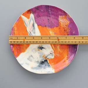 May include: A decorative ceramic plate with a white rabbit illustration set against an orange and purple background. The plate is approximately 23 cm in diameter, with a painted design and a white rim.