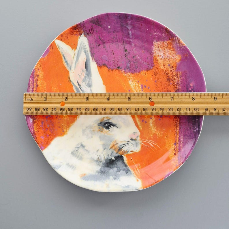 May include: A decorative ceramic plate with a white rabbit illustration set against an orange and purple background. The plate is approximately 23 cm in diameter, with a painted design and a white rim.