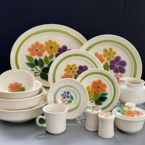 May include: A vintage floral dinnerware set featuring plates, bowls, a creamer, and a covered dish. The off-white pieces have a green rim and are decorated with orange, yellow, and purple flowers. A matching mug and salt and pepper shakers are also included.