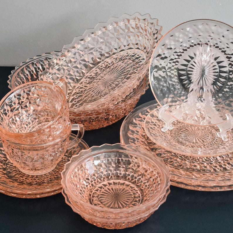 K&ouml;nnte beinhalten: Eine Sammlung rosafarbener Depression-Glasgeschirr, darunter Teller, Schalen und Tassen mit Untertassen. Das Glasgeschirr weist ein Diamantmuster und gewellte Kanten auf. Das Set ist auf einer dunklen Oberfl&auml;che angeordnet.