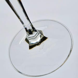 K&ouml;nnte beinhalten: Nahaufnahme von Fu&szlig; und Stiel eines transparenten Weinglases. Der Stiel hat ein facettiertes Design, und der Fu&szlig; ist rund. Das Glas ist transparent, das Licht reflektiert sich auf den Oberfl&auml;chen. Das Bild ist vor wei&szlig;em Hintergrund.