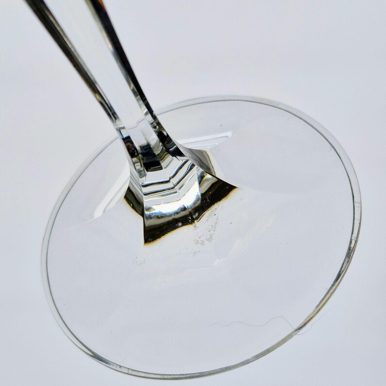 K&ouml;nnte beinhalten: Nahaufnahme von Fu&szlig; und Stiel eines transparenten Weinglases. Der Stiel hat ein facettiertes Design, und der Fu&szlig; ist rund. Das Glas ist transparent, das Licht reflektiert sich auf den Oberfl&auml;chen. Das Bild ist vor wei&szlig;em Hintergrund.