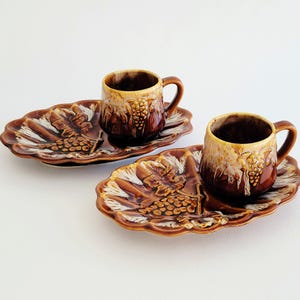 May include: Two-piece ceramic set featuring a cup and a matching tray. The set is brown with a grape design and a white accent. The cup has a handle and the tray is oval-shaped. The set is designed for serving coffee or tea.