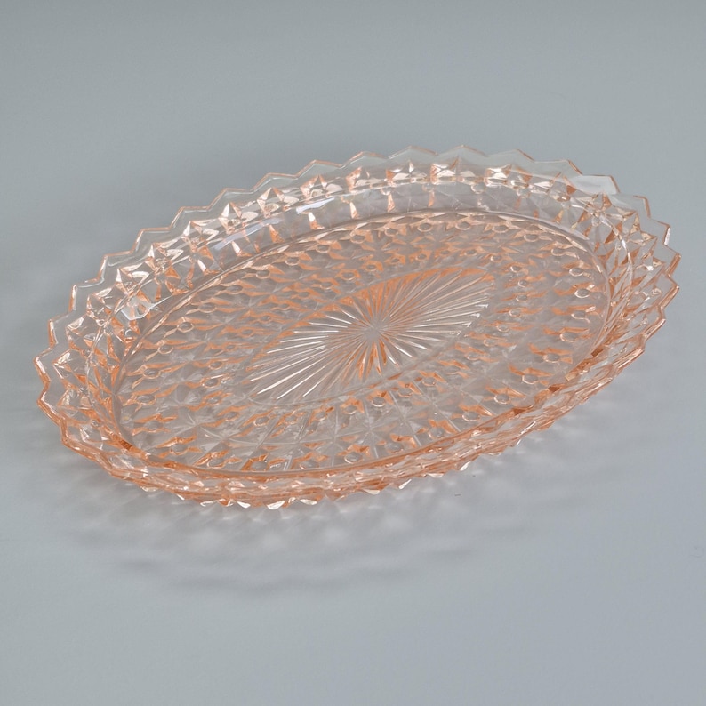 K&ouml;nnte beinhalten: Ein ovales Serviertablett aus rosa Glas. Das Tablett hat eine strukturierte Oberfl&auml;che und einen dekorativen Zickzackrand. Die Mitte des Tabletts hat ein Sternmuster. Dieses Vintage-St&uuml;ck ist ideal zum Servieren von Vorspeisen oder Desserts, ca. 30 cm lang.