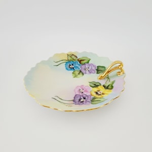 May include: A white porcelain serving dish with a gold rim and handle. The dish is decorated with hand-painted purple, yellow, and blue pansies.