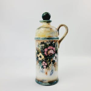 May include: A ceramic pitcher with a floral design. The pitcher is decorated with a bouquet of pink, white, and yellow flowers, green leaves, and berries. The pitcher has a green lid and a handle.