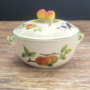 May include: A white ceramic serving bowl with a lid, featuring a green rim and handles. The lid is topped with a peach-shaped handle. The bowl is decorated with illustrations of apples, leaves, and berries. The bowl sits on a wooden surface.