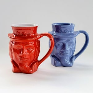May include: Two ceramic mugs with a face design. One mug is red with a white interior, and the other is blue. Both mugs have a handle and a hat-shaped top.